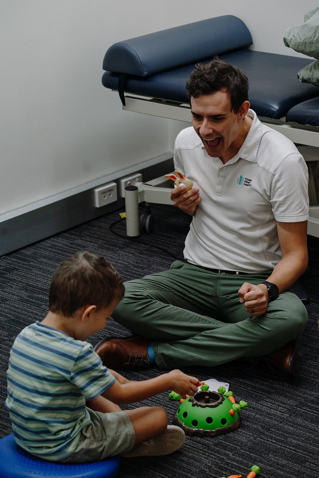 Speech pathologist Jonathon Cronk laughing as he holds up a toy rabbit for a young boy during a floor session