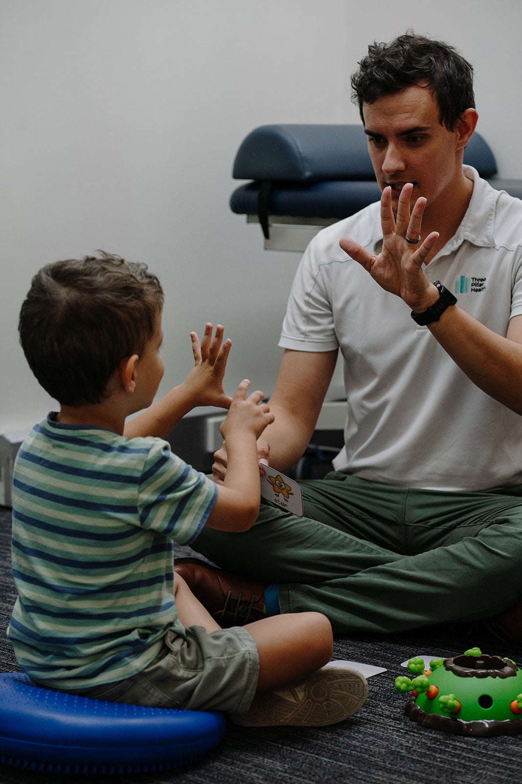 Speech pathologist Jonathon Cronk and a young boy each holding a hand up during a session on the floor